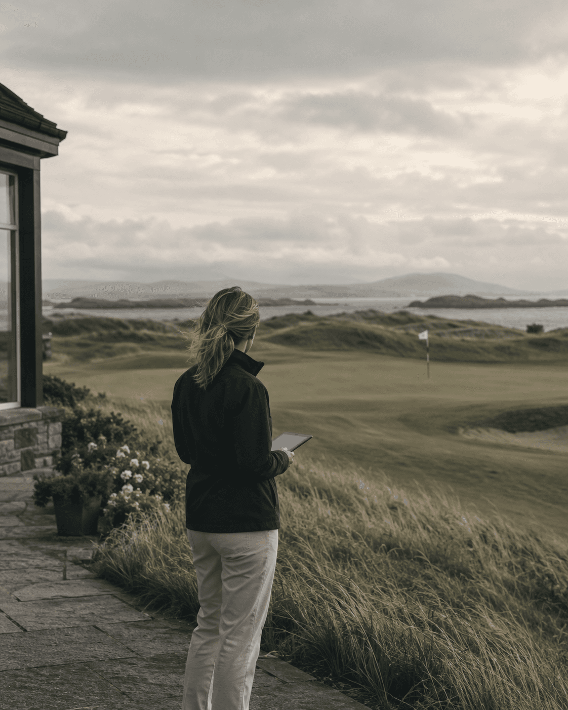 A general manager with a tablet looks out over a links course from the clubhouse terrace.